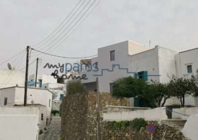 White-washed Mediterranean street with blue-trimmed buildings, a winding cobblestone path, and a stone retaining wall under an overcast sky, with overhead power lines and a no-parking sign in the foreground.